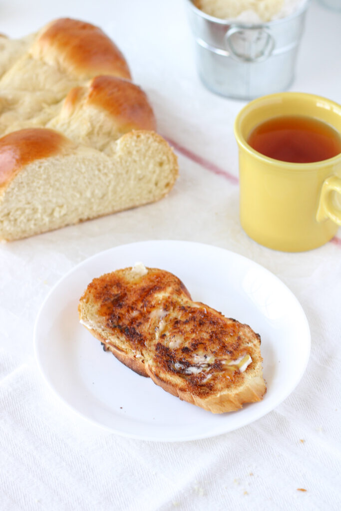 Slice of challah, toasted with butter and honey on a plate. Loaf and mug of tea in the background.