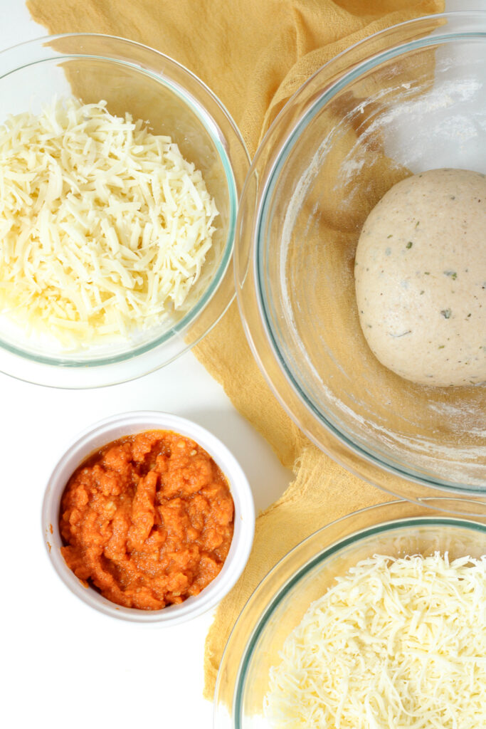 Two bowls of shredded cheese, one bowl of pizza dough, and a ramekin of pumpkin puree
